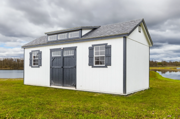 White storage shed with gray shutters, dormer windows, and shingle roof